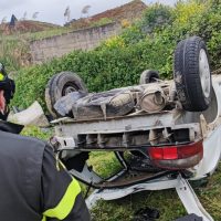 Auto ribaltata su un lato in mezzo a erba alta, con ruote in alto e danni su cofano e paraurti; operatore di soccorso in primo piano.