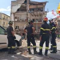 Quattro vigili del fuoco in abiti completi osservano un edificio parzialmente crollato in una zona urbana, durante un intervento di soccorso.