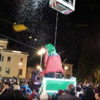 Grande figura in costumi colorati sorregge un pallone illuminato, festa notturna con folla e edifici sullo sfondo; atmosfera festosa.