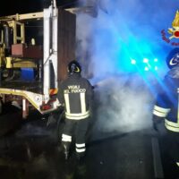 Vigili del fuoco in azione durante un incidente notturno, fumo visibile e lampeggianti blu, operano vicino all’accessorio di un veicolo d’emergenza.