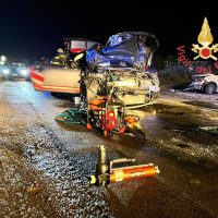 Incidente stradale di notte: auto e scooter distrutti, strumenti e schematiche sul selciato, sirene coperte da luci rosse.