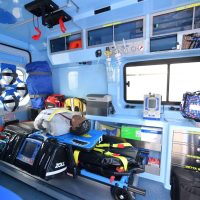 Interno di un'ambulanza attrezzata: apparecchiature mediche, zaini, contenitori e schermi pronti per soccorso, in colori blu e bianco.