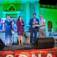Quattro persone sul palco durante un evento, luci verdi e una cantante a sinistra, pubblico sullo sfondo.
