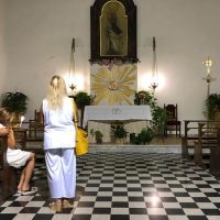 Due donne pregano davanti all'altare in una chiesa luminosa, pavimento a scacchi e decorazioni floreali dorate.