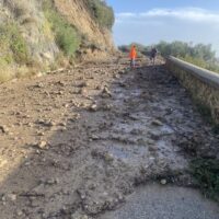 Strada sterrata discontinua e polverosa sul costone, con massi e terra, guard-rail a destra e operai al lavoro in fondo.