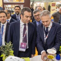 Due imprenditori al tavolo in fiera, mentre scambiano note sul prodotto tra visitatori sorridenti e vetrine illuminate sullo sfondo.