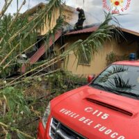 Auto dei vigili del fuoco rossa in primo piano, edificio sfondato e tetto gravemente danneggiato, piante cadenti, situazione di emergenza.