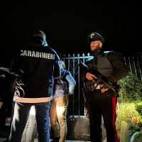 due Carabinieri armati in divisa scura controllano una persona all’ingresso di una zona recintata durante la notte