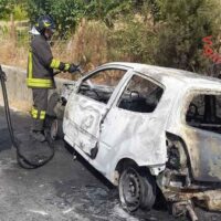 Fiammeggianti operazioni di spegnimento: vigile del fuoco spegne un’auto incendiata sul lato strada, fumo e danni visibili ai veicoli.