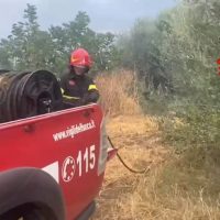 Pompiere in tuta rossa sul bordo di una jeep fuoristrada, fumo e vegetazione secca sullo sfondo, scena di intervento antincendio.