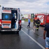 Ambulanza e veicoli di soccorso su strada, equipaggi in intervento su un incidente stradale, persone presenti sul lato della carreggiata.