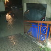 Strada allagata di notte, pedane e riflessi sull’acqua, balcone con ringhiera e contenitore blu posizionato accanto a una rampa.