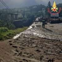 Strada fangosa e macchia d’acqua lungo una singolare via, con mezzi e persone all’orizzonte, vignetta di protezione civile sul lato superiore.