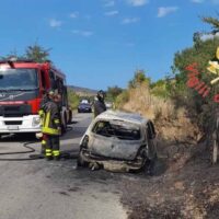 Automobile distrutta ai margini della strada, vigili del fuoco e camion dei soccorsi presenti; contesto stradale con vegetazione secca e cielo sereno.