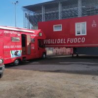 Auto del reparto Vigili del Fuoco rosso, mezzi di soccorso parcheggiati, edificio moderno sullo sfondo, giornata soleggiata.