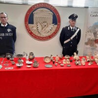Due ufficiali e una quantità di oggetti d’epoca e medaglie disposti su tavolo rosso, allestimento ufficiale dell’Arma dei Carabinieri.