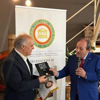 Due uomini in abiti formali si scambiano un libro durante un evento, con banner istituzionale sullo sfondo.