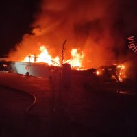 Incendio notturno in un parcheggio: fiamme arancioni che avvolgono veicoli e pali, fumo intenso e atmosfera di emergenza.