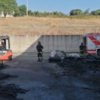 Azienda di soccorso: due vigili del fuoco e un veicolo di emergenza su fondale asfalto scuro, rogo spento vicino a una scarpata erbosa.