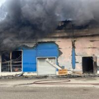 Edificio in fiamme con tedieri di fumo nero; vetri infranti, persiane blu, veicoli e cavi sul marciapiede, scena urbana devastata.