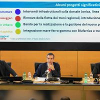 Uomini in una conferenza seduti a un tavolo, sfondo di presentazione con testo in italiano e grafici, ambiente formale.