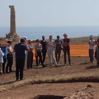 Gruppo di persone in un cantiere o sito archeologico vicino al mare, con palizzate arancioni e una colonna rinoceronte, sullo sfondo l’acqua.