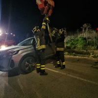 Squadra di vigili del fuoco al lavoro in strada notturna, con autovettura incidentata e pancate di luci d’emergenza.