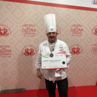 Chef italiano in camicia bianca e cappello da cuoco, posa con diploma su sfondo decorato a tema cucina italiana.