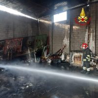 Vigili del fuoco in una rimessa danneggiata dal fuoco, intervento con idrante e fumo visibile nell’ambiente.