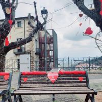 Panchina in cortile con cuori rossi appesi agli alberi spogli, sfondo urbano e cielo parzialmente nuvoloso
