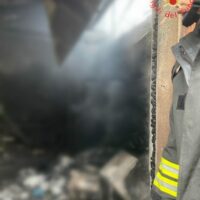 Operatore in tuta riflettente osserva un baccello di acciaio e fumo sullo sfondo, primo piano sfocato, scena di cantiere industriale.