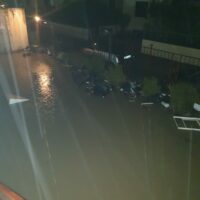Veduta notturna di una strada bagnata con luci riflettenti sull’acqua, autovetture parcheggiate e un’area industriale sullo sfondo.
