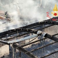 Taglio di una struttura in legno carbonizzata tra fumo bianco e roghi, resti incendiati e scheletri di travi.