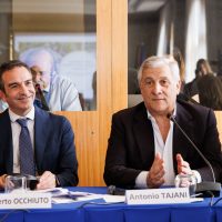Due uomini seduti a tavola durante una conferenza, uno sorridente e l’altro parlante, con nomi su placard sul tavolo.