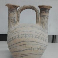 Antica brocca in ceramica a forma di orecchio con impugnature doppie, decorazioni a righe azzurrine, sfondo bianco.