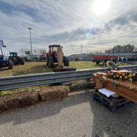 Immagine di una zona industriale con mezzi pesanti, trattori e carichi su pallets vicino a una barriera metallica e container, cielo sereno.