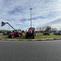 Trattori agricoli in fila su prato vicino a strada, con accessori e cielo sereno, scena rurale italiana.
