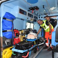 Passeggeri e soccorritori in ambulanza: sedato paziente in barella con supporti, operatori in divise high visibility e attrezzature mediche a bordo.