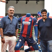 Due uomini in piedi accanto a una mascotte in uniforme da calcio rosso e blu, in un evento sportivo all'aperto.
