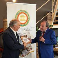 Due uomini in abiti formali si scambiano un premio al momento di una cerimonia, con banner istituzionale sullo sfondo.