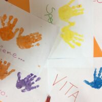 Mani colorate in vernice su carta bianca con scritte in italiano, angoli arancioni, design giocoso e vivace.