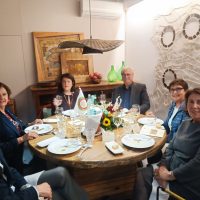 Gruppo di persone sedute attorno a un tavolo rotondo: cibo, caffè e fiori, atmosfera conviviale in una sala accogliente.