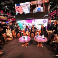 Panel discussion in uno spazio espositivo con pubblico, tavolini rotondi e schermi grandi alle spalle, atmosfera vivace e professionale.