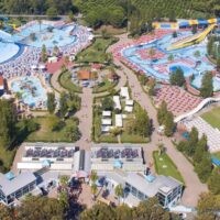 Panoramica di un parco acquatico dall’alto: piscine a olio d’acqua, scivoli colorati, lettini e vialetti tra verde e strutture ben illuminate.