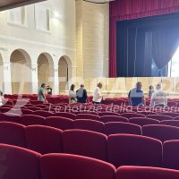 Interno di un teatro con palcoscenico, tendaggi viola e file di sale rosse; persone in sala discutono tra teatri di legno e archi.