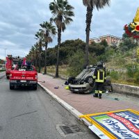 Incidente su strada: vigili del fuoco e soccorritori vicino a un’auto rovesciata, SUV giallo davanti, scena su carreggiata con cartelloni e palme sullo sfondo.