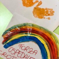 Arcobaleno dipinto a tempera con impronte di mani, scritte italiane su sfondo bianco e dettagli verdi.