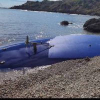 Scafo di una barca incagliata su una spiaggia rocciosa, con acque calme e promontori sullo sfondo.