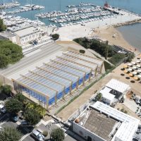 Veduta aerea di marina e spiaggia: banchi e strutture ombreggiate, ormeggi, e il mare sullo sfondo in una zona balneare italiana.