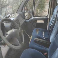 Interni di un autocarro: volante, cruscotto e sedili blu, finestrini laterali e vista esterna di alberi.